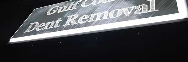 The illuminated 'Gulf Coast Dent Removal' shop sign at night, the brushed-metal panel catching the light